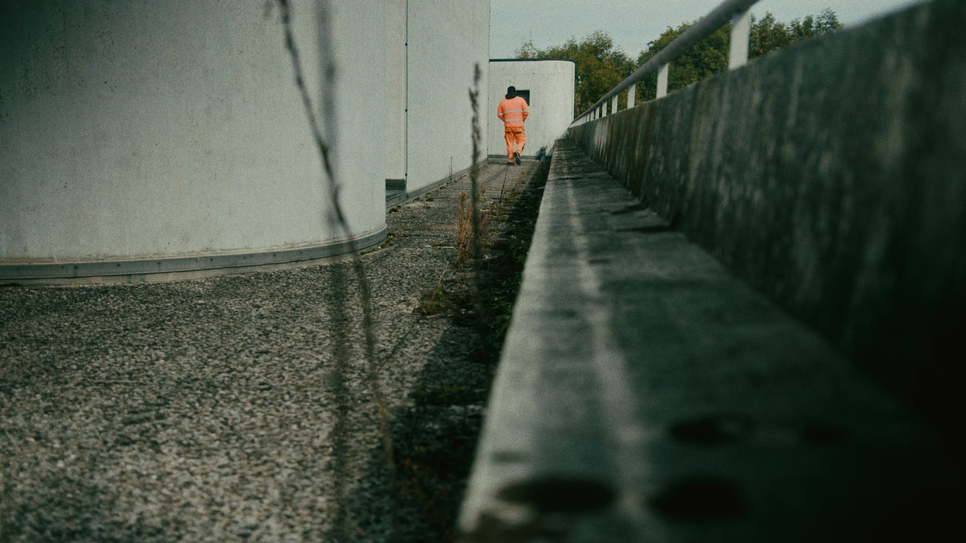 Eine Person in orangefarbener Arbeitskleidung geht durch einen schmalen Betonweg.